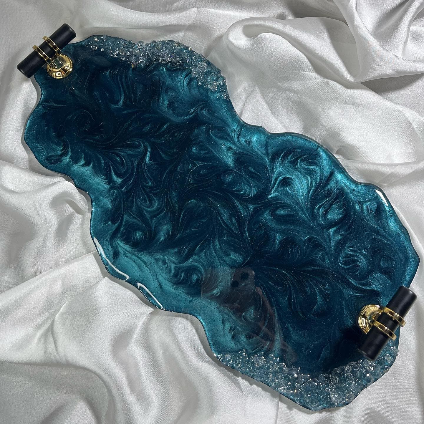 Wave-edged teal resin serving tray with a swirled pearlescent surface, clear crystal chips along two edges and black and gold handles